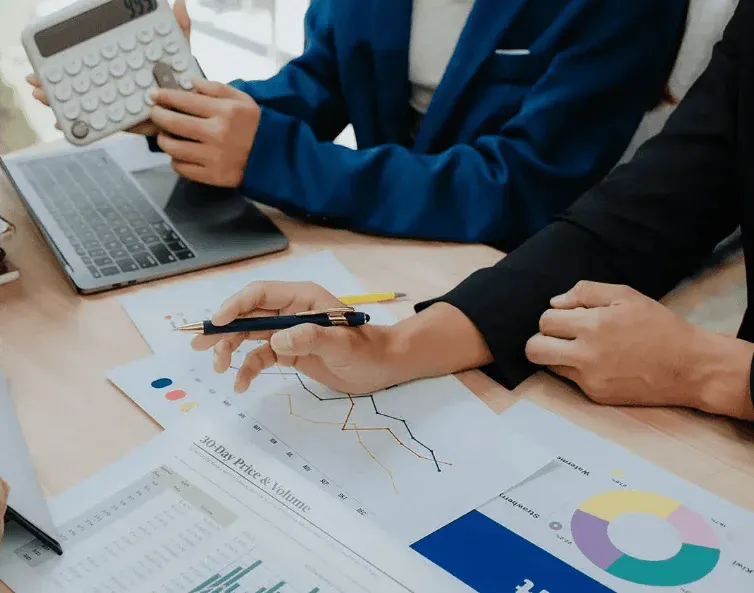 Equipe analisando gráficos e planilhas em uma reunião de negócios, com uma pessoa usando um laptop e anotando dados em documentos com charts e indicadores.