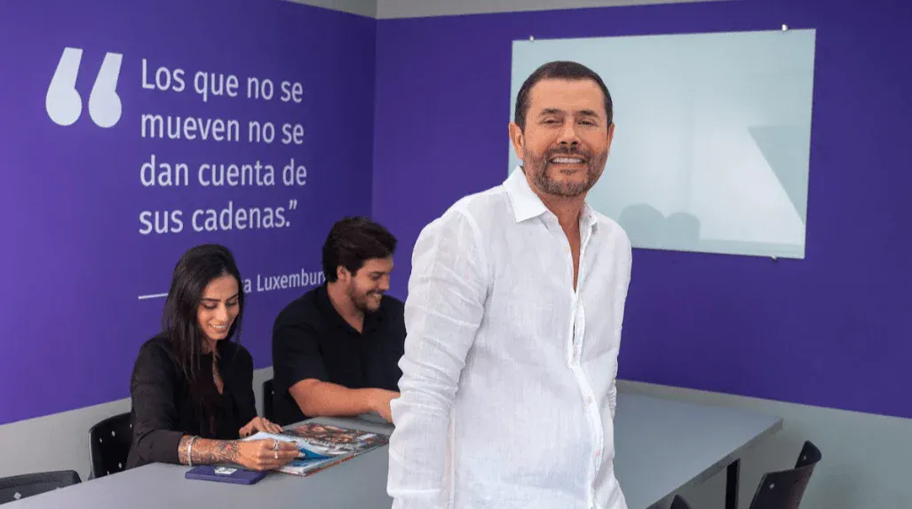 Homem de camisa branca em apresentação ao lado de outras pessoas sentadas em mesa com material impresso, em um ambiente com fundo roxo e citação motivacional ao lado.