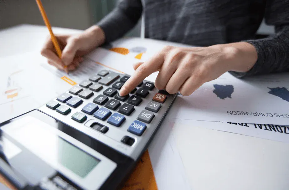 Pessoa digitando códigos em um teclado de computador financeiro ao usar uma calculadora, com planilhas e documentos ao fundo, em ambiente de trabalho.