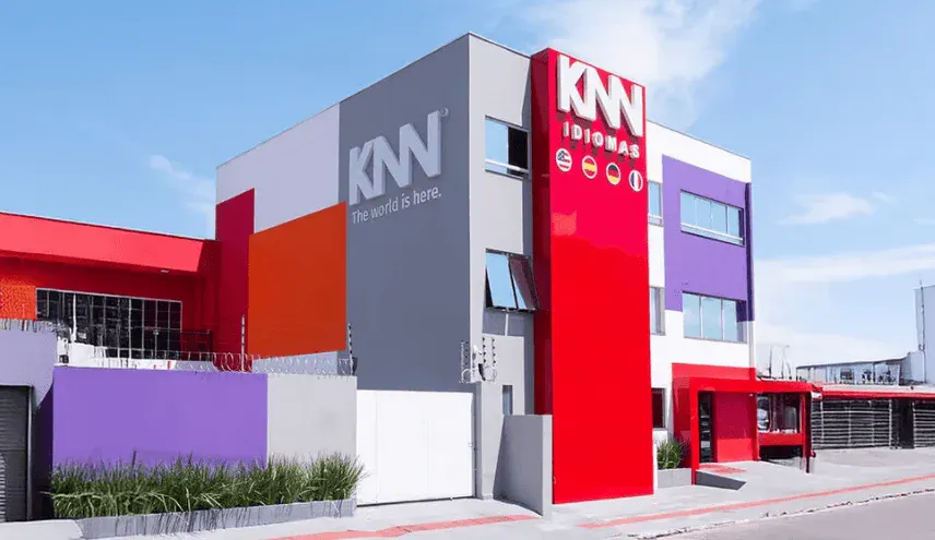 Fachada moderna do prédio da KNN Idiomas, com letras e logo em destaque, paredes coloridas e janelas amplas em ambiente externo com céu azul ao fundo.