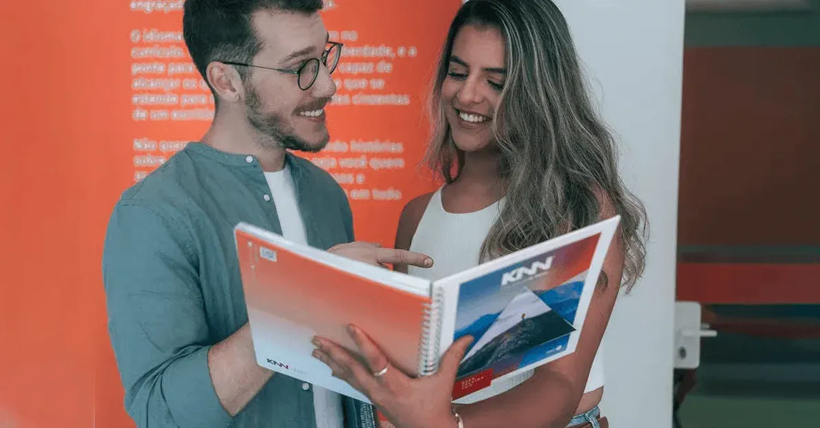 Dois jovens em um ambiente de evento, sorrindo e consultando um folder com a marca KMV, enquanto conversam. Foto em destaque para comunicação e atendimento.