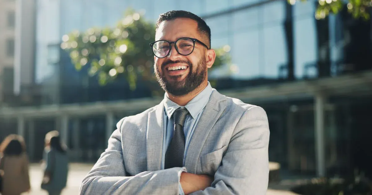 Homem de terno e gravata, com barba e óculos, sorrindo e cruzando os braços em ambiente corporativo ao ar livre, com prédios ao fundo.