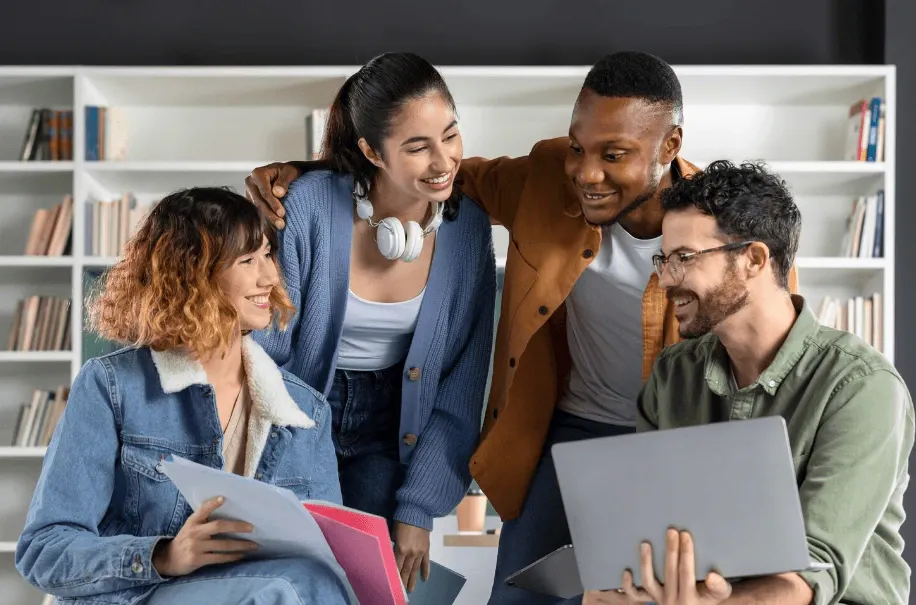 Grupo de amigos em ambiente de escritório ou sala de estudo, sorrindo e compartilhando informações em computadores e tablets, com uma pessoa usando fones e os outros lendo documentos