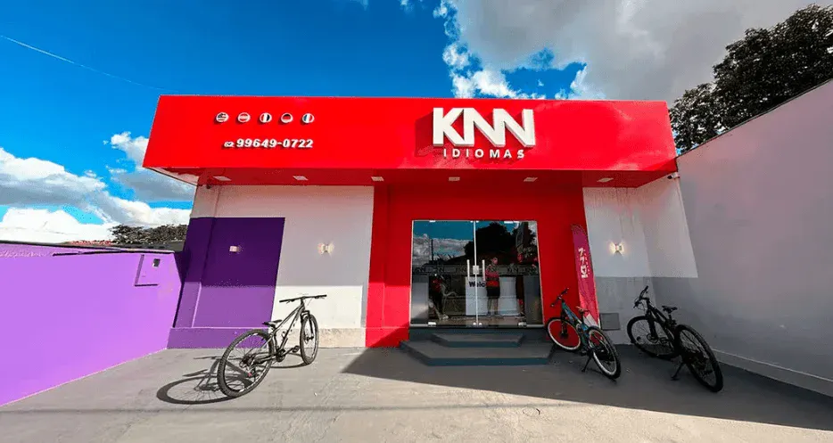 Fachada de uma escola de idiomas com a placa KN Idiomas em destaque, bandeja vermelha, bicicletas estacionadas e céu azul ao fundo.