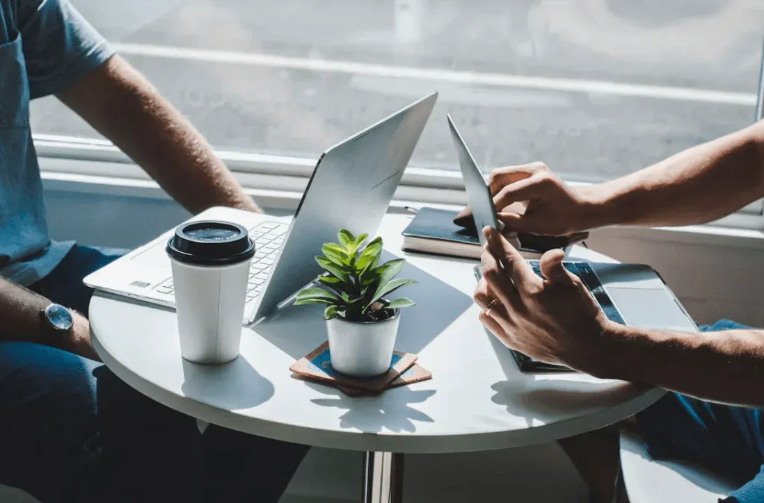 Três pessoas trabalhando em uma mesa circular ao ar livre, com notebooks, caneca térmica e um vaso de planta ao centro enquanto uma delas segura um tablet e outra usa o laptop