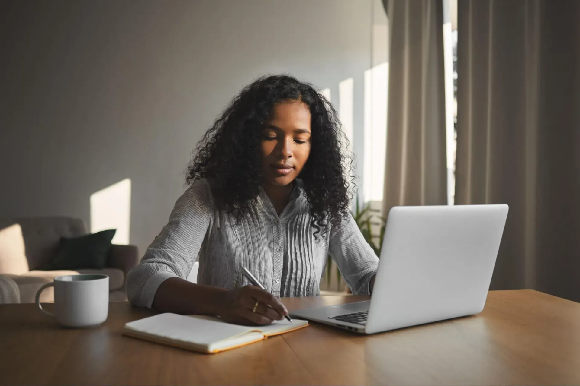 Mulher com cabelo cacheado trabalhando em um escritório em casa, escrevendo em um caderno e usando notebook sobre uma mesa com xícara de café ao lado, em luz natural.