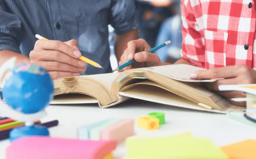 Crianças e adultos desenhando e escrevendo em uma mesa, com lápis coloridos e um livro aberto, em atividade educativa de sala de aula ou ateliê.