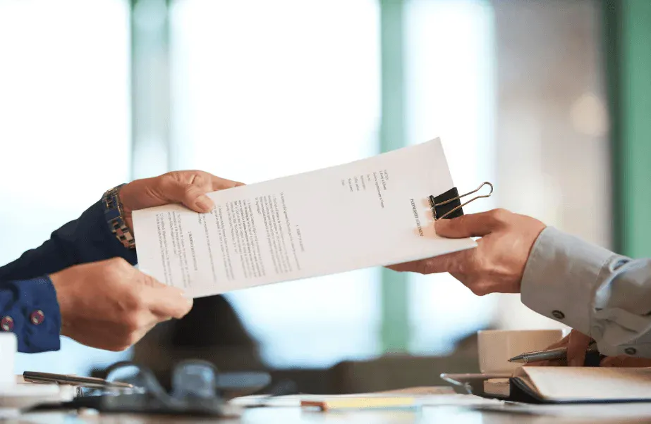 Duas pessoas em ambiente corporativo assinam um documento, com as mãos segurando uma folha impressa sobre uma mesa de trabalho, sugerindo formalização de contrato.