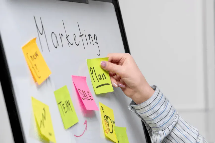 Pessoa marcando post-its coloridos em quadro branco com anotações “Marketing” e “Plan”, indicando planejamento e organização de tarefas.