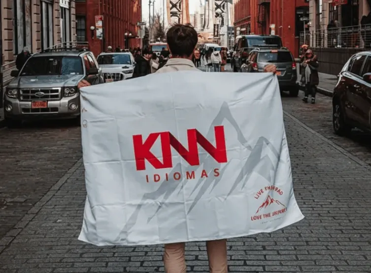Pessoa em uma rua movimentada com carros e prédios ao fundo, usando uma bandeira/cover branca com a marca KNN IDIOMAS visível.