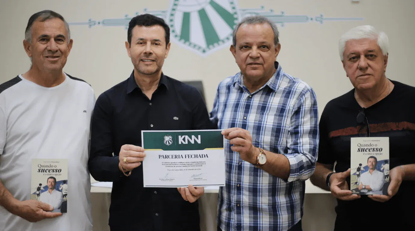 Homens em evento empresarial seguram certificados de parceria e livros, com logotipo KNN visível na placa e fundo com banner na parede.