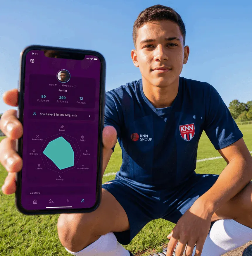 Jovem no campo de futebol, sorrindo e segurando um celular com aplicativo de treinamento exibindo ranking, seguidores, badges e métricas de desempenho