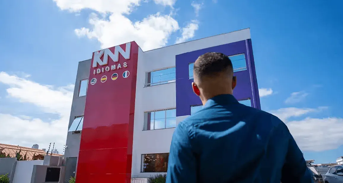 Vista externa de uma unidade da KNN IDIOMAS, com fachada vermelha e branca e letreiro KM DIOMAS, enquanto uma pessoa de costas observa o prédio sob céu azul com nuvens.