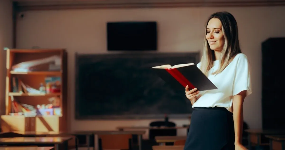 Mulher em um escritório ou sala de estudos, lendo um livro aberto com expressão serena, ao lado de mesa com materiais e ao fundo quadro e equipamentos, em ambiente interno.