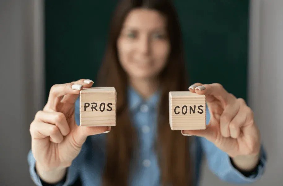 Imagem desfocada de uma mulher segurando dois blocos de madeira com as palavras “PROS” e “CONS”, representando prós e contras de uma decisão.