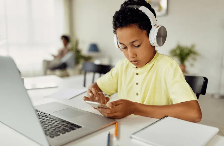 Criança usando fones de ouvido grandes e estudando no computador em uma mesa, com foco na tela e no celular, em ambiente doméstico claro