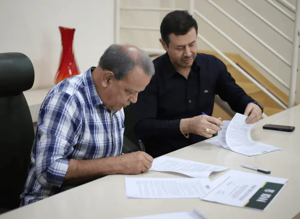 Dois homens assinando documentos em uma mesa, em ambiente interno com escadas ao fundo, enquanto analisam papéis e caneta durante um processo formal
