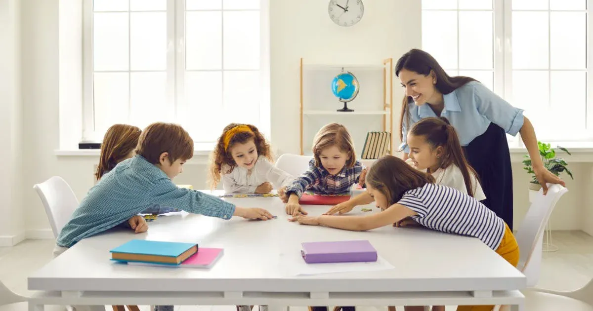 Professora sorri enquanto ensina crianças ao redor de uma mesa com cadernos e livros, em sala de aula iluminada com janelas grandes e decoração ao fundo.