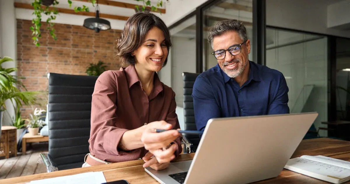 Duas pessoas sorrindo em uma sala moderna, analisando documentos e usando notebook durante uma reunião ou trabalho em equipe, com um tablet na mesa