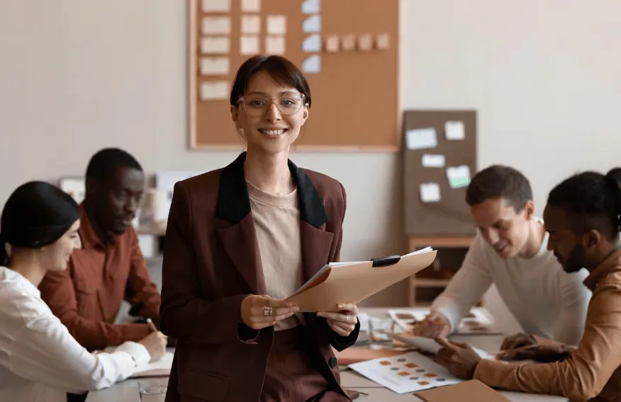 Mulher executiva sorridente apresenta documentos e lidera reunião em equipe, em ambiente corporativo com pessoas analisando gráficos e trabalhando em mesa, foco no trabalho colaborativo.
