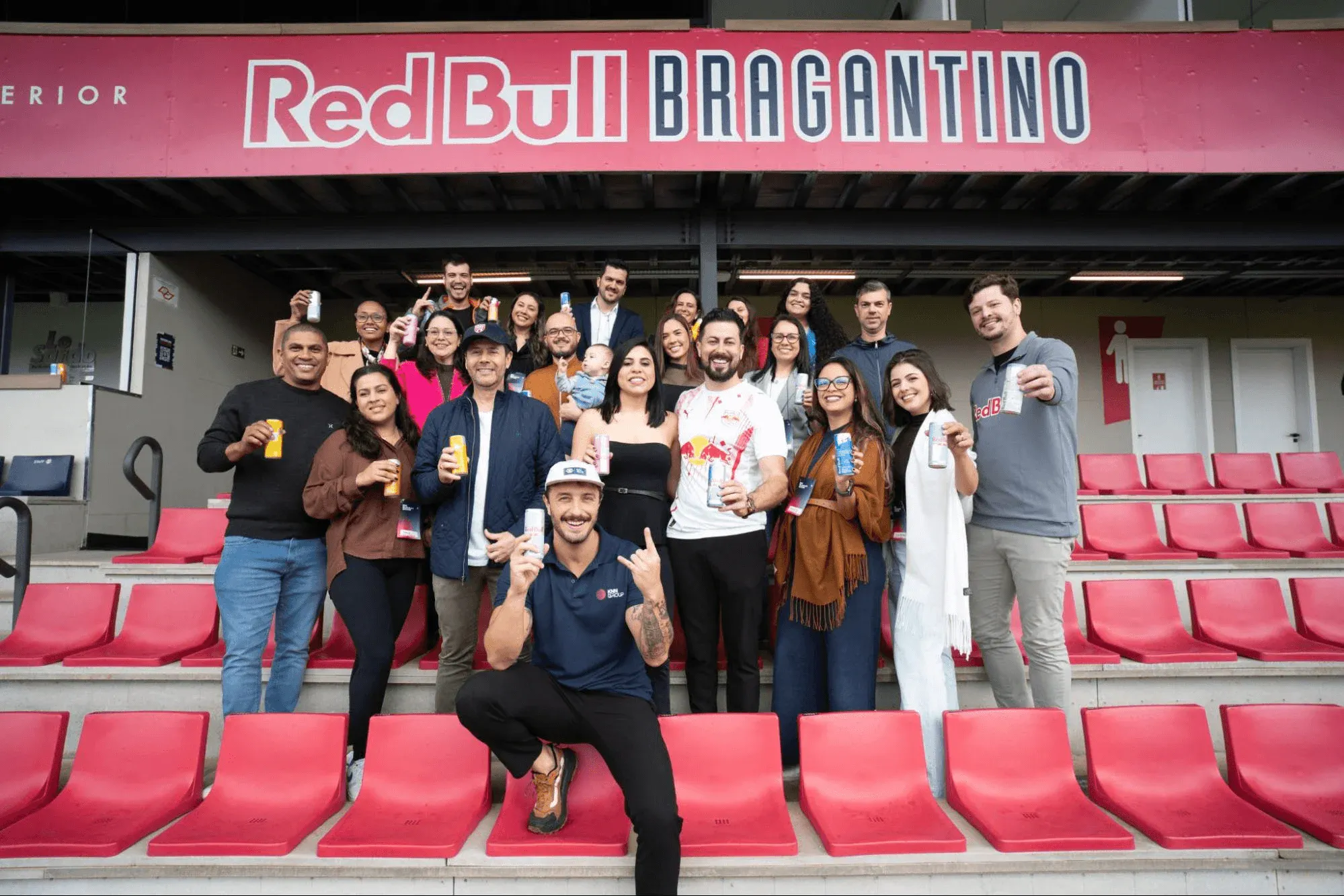 Grupo de pessoas posando para foto em estádio de futebol, com cadeiras vermelhas e faixa grande escrito “Red Bull BRAGANTINO” ao fundo.