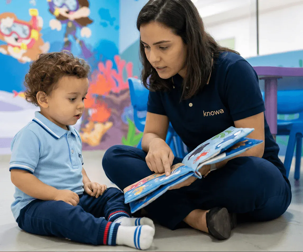 Professora da Knowa sentada no chão com uma criança em sala de aula infantil, mostrando um livro ilustrado e participando de uma atividade de leitura e aprendizado.
