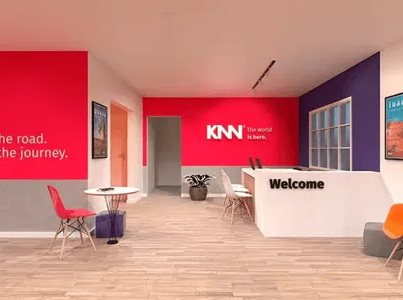 Interior de um lounge moderno com paredes coloridas em vermelho e roxo, recepção com placa “Welcome”, mesa de apoio e cadeiras, com logo KNN ao fundo.