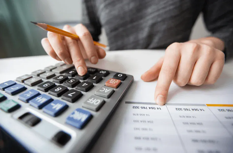 Pessoa usando calculadora e caneta para revisar números e contas em uma mesa de trabalho, com documentos ao fundo e atenção ao cálculo financeiro.