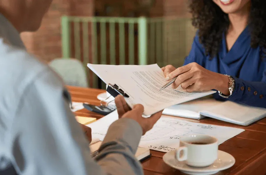 Encontro de negócios em um escritório: duas pessoas analisam e assinam documentos sobre uma mesa, com caneta, folhas e xícara de café ao fundo.