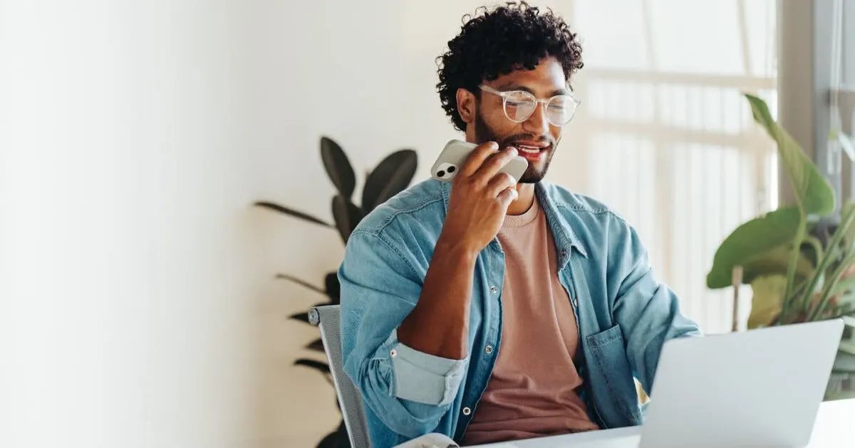 Homem jovem com óculos e cabelo cacheado falando ao celular enquanto trabalha em um computador, sentado em um ambiente de escritório moderno e iluminado com plantas ao fundo.