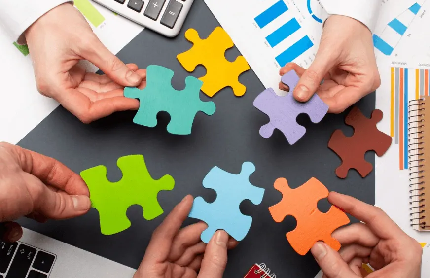 Mãos encaixando peças de quebra-cabeça coloridas sobre uma mesa, simbolizando colaboração e trabalho em equipe com foco em estratégia e integração.