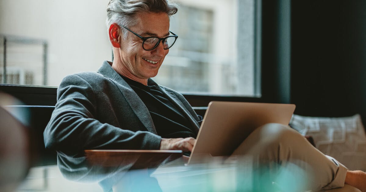 Homem de óculos sentado à mesa com um laptop, concentrado em seu trabalho.