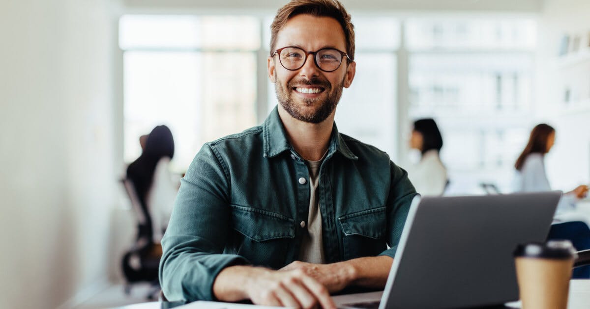 Homem sorridente com óculos sentado à mesa, utilizando um laptop.