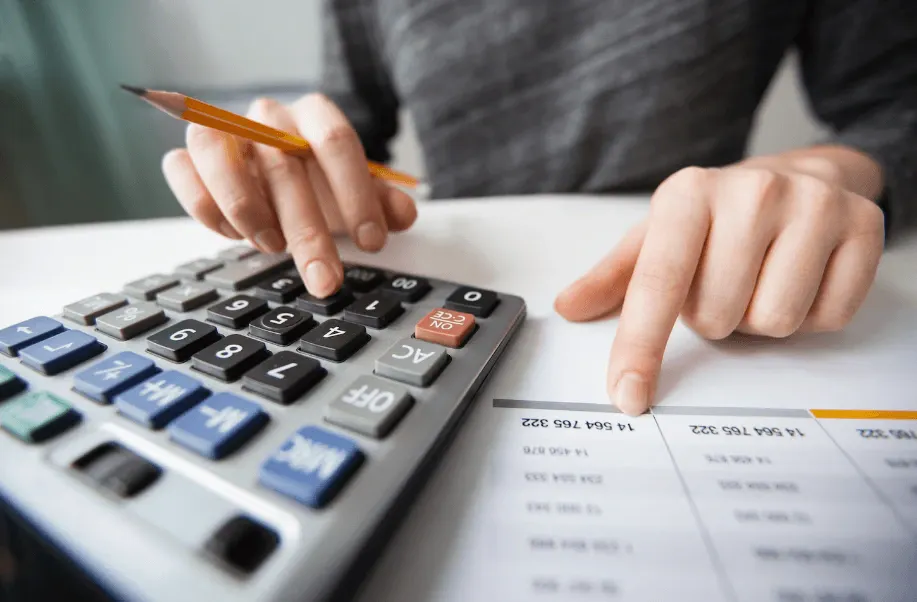 Pessoa usando calculadora com caneta e analisando números e valores em planilha, indicando controle financeiro e cálculos de orçamento.
