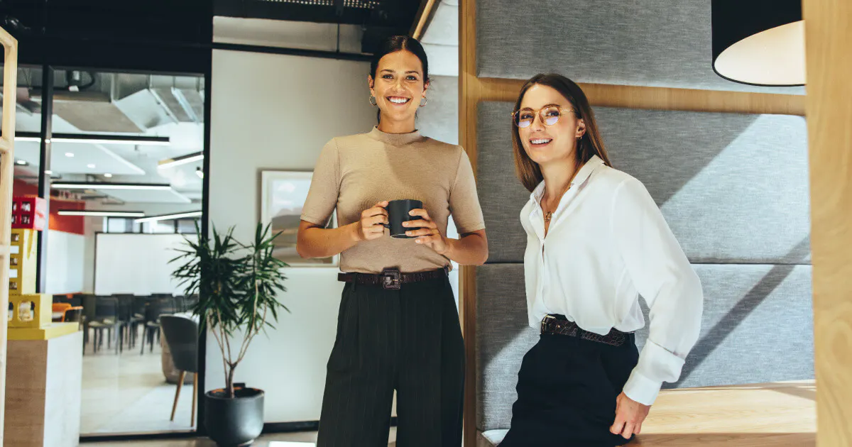 Duas mulheres em um escritório, uma segurando uma xícara de café, conversando e sorrindo.