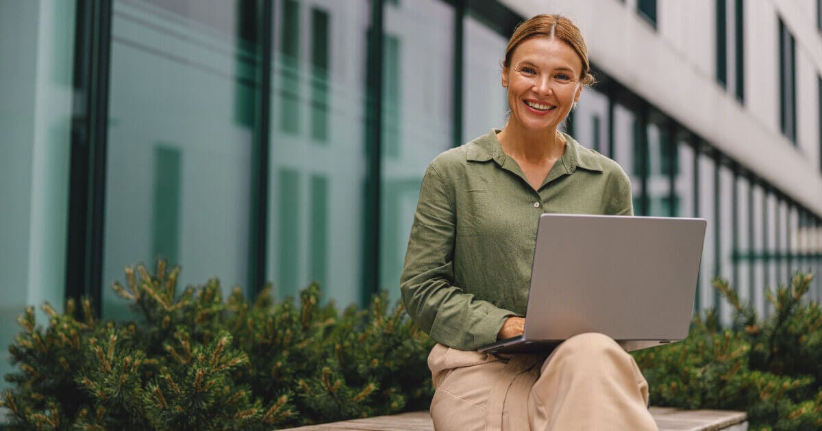 Mulher sentada em um banco, utilizando um laptop, em um ambiente ao ar livre.