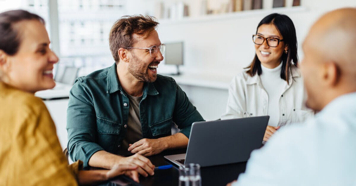 Três pessoas sentadas à mesa, utilizando um laptop, em um ambiente de trabalho colaborativo.