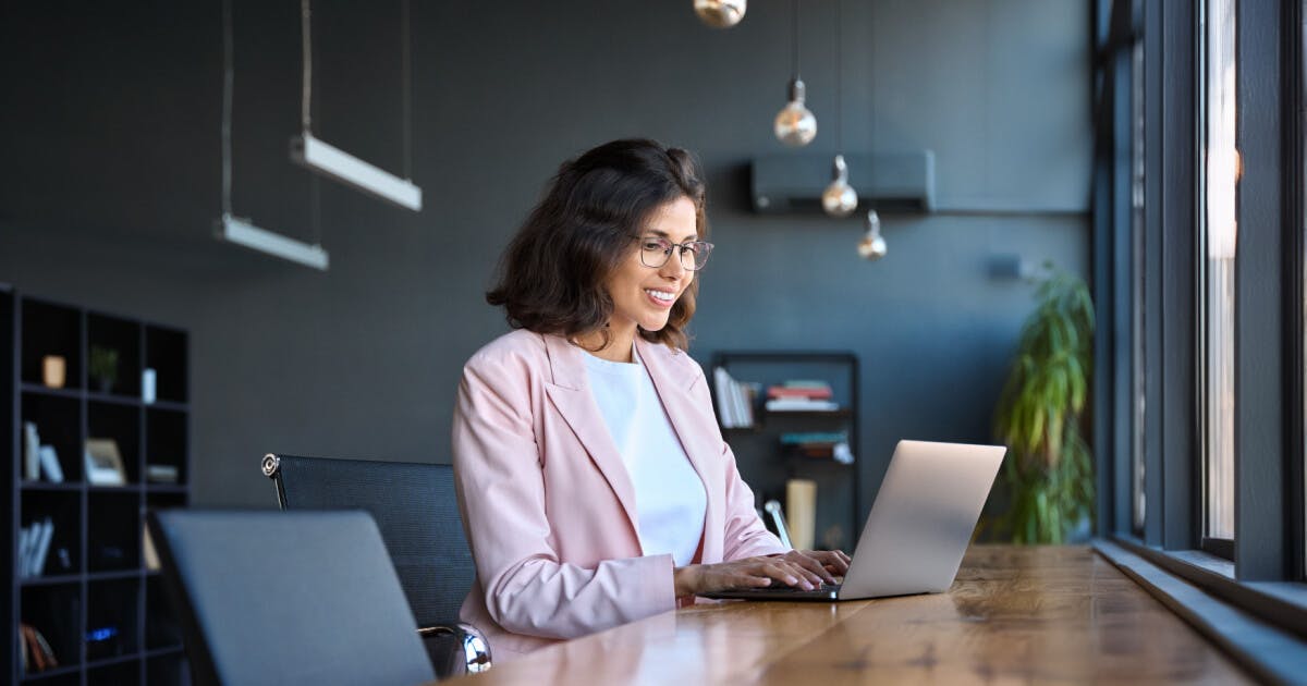 Mulher com blazer rosa sentada à mesa, utilizando um laptop.