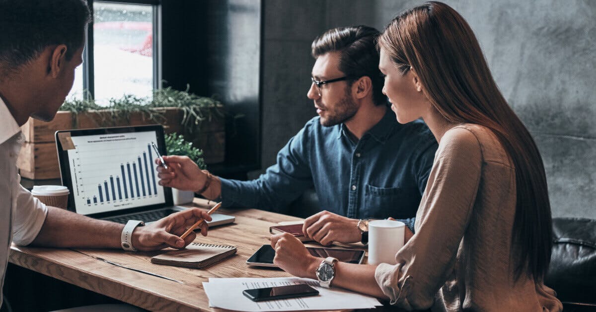 Três pessoas sentadas à mesa, utilizando um laptop e cercadas por papéis, em um ambiente de trabalho colaborativo.