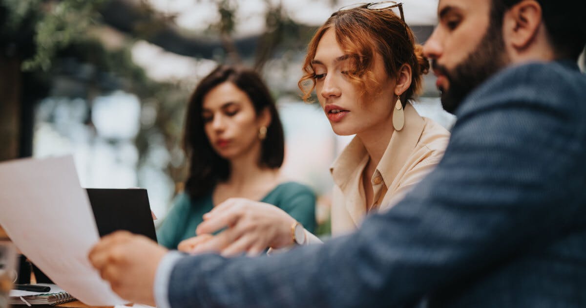 Três pessoas sentadas à mesa, analisando documentos e discutindo em um ambiente colaborativo.