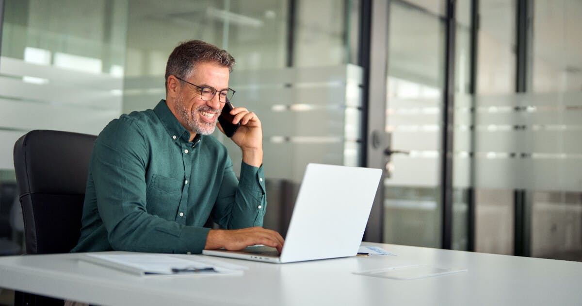 Homem sentado à mesa com um laptop, conversando ao telefone, em um ambiente de trabalho.