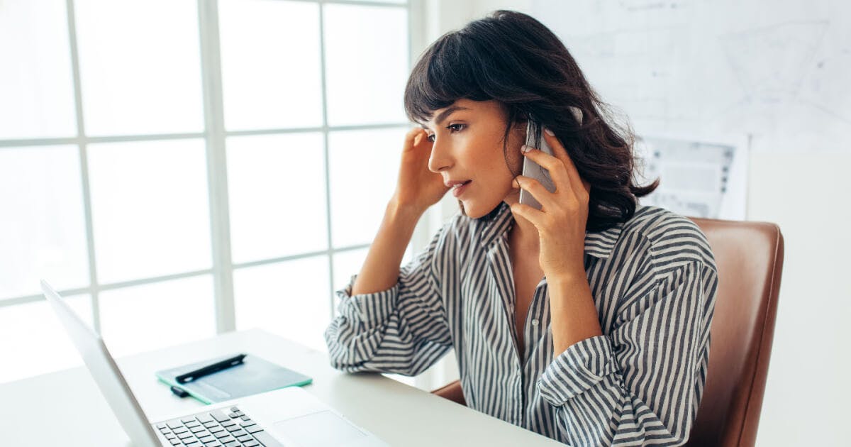 Mulher sentada à mesa, apoiando a cabeça no telefone, demonstrando concentração e envolvimento na conversa.