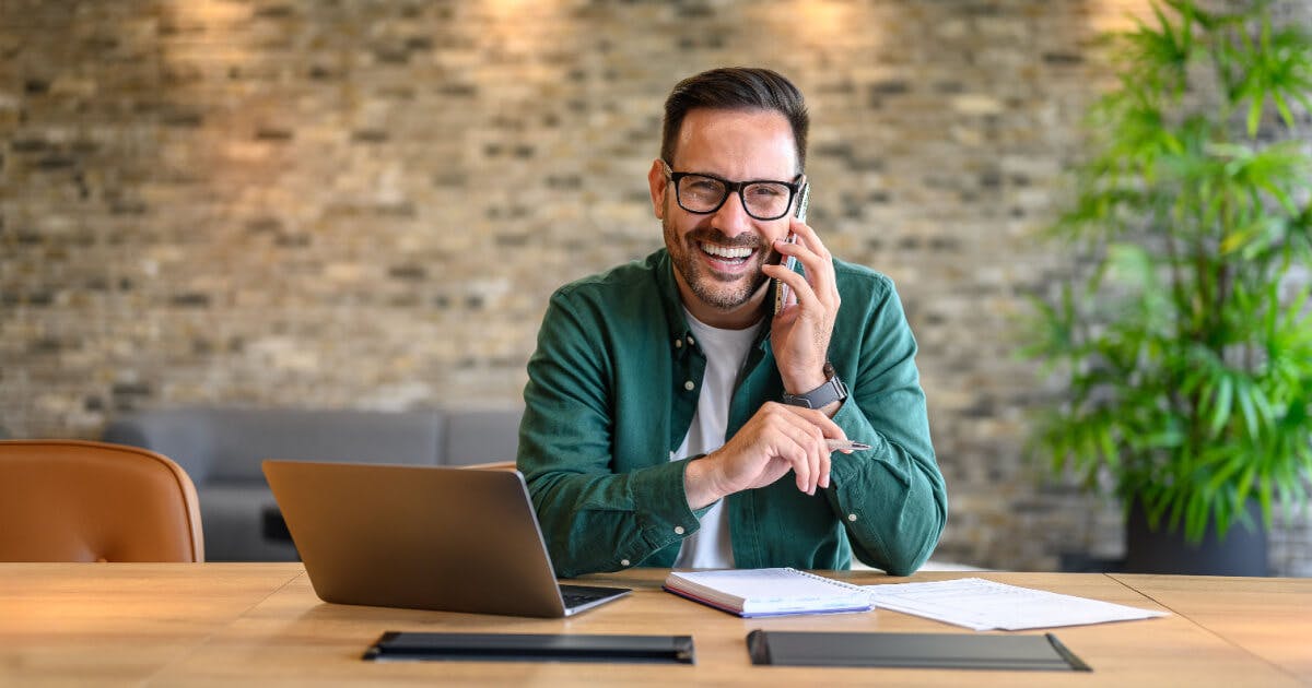 Homem de óculos sentado à mesa, utilizando um laptop e um telefone.