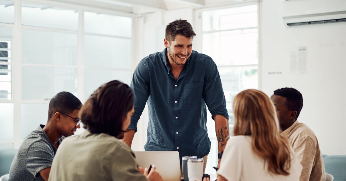 Um homem de pé orientando dois homens e duas mulheres que estão sentados em uma mesa.