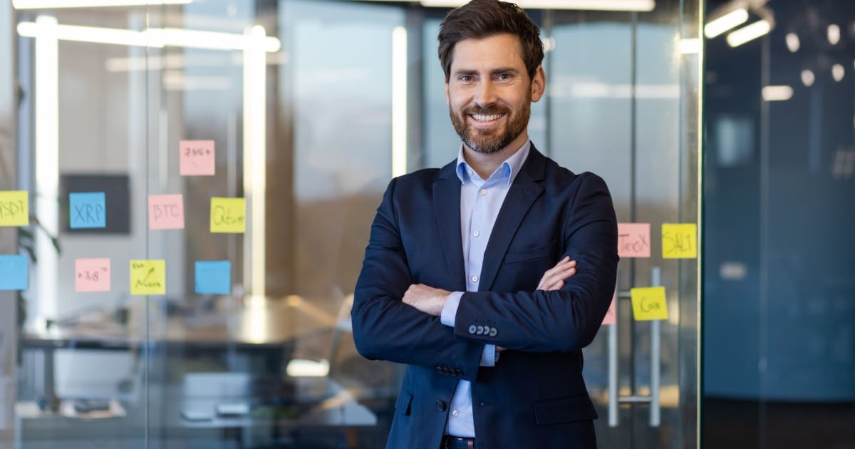 Um homem de barba com terno e roupa social de braços cruzados e sorrindo.