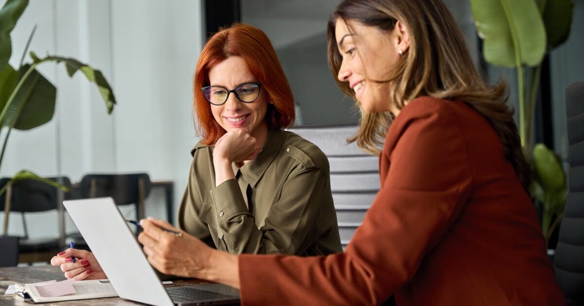 Duas mulheres sentadas à mesa, utilizando um laptop, em um ambiente de trabalho colaborativo.