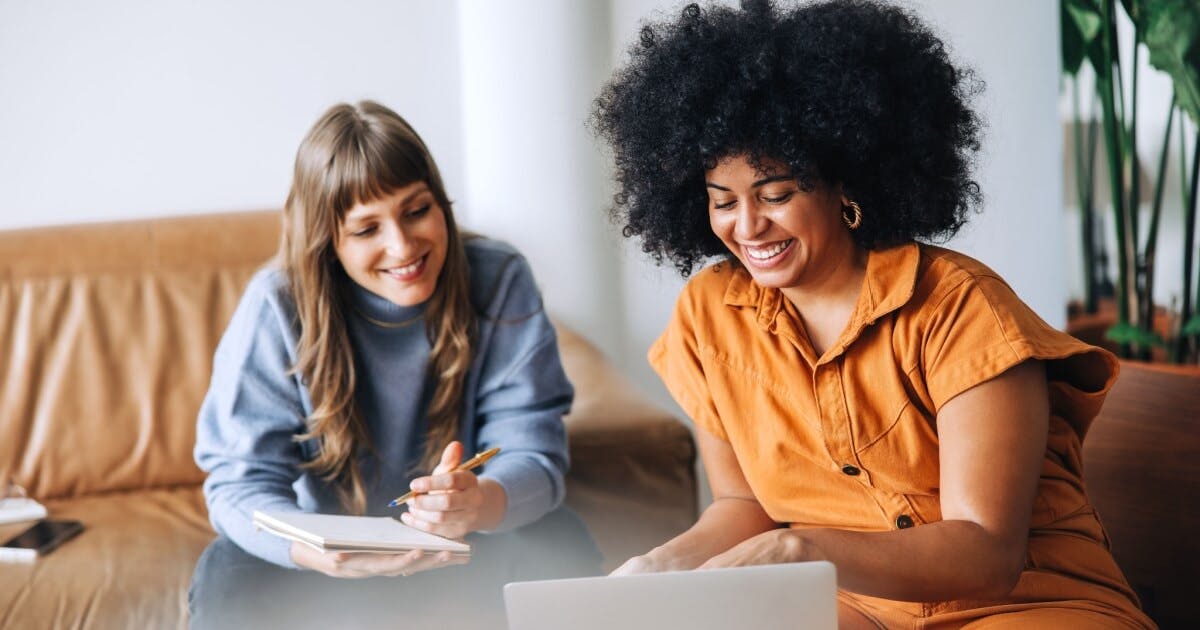 Duas mulheres sentadas em um sofá, utilizando um laptop, em um ambiente aconchegante e colaborativo.