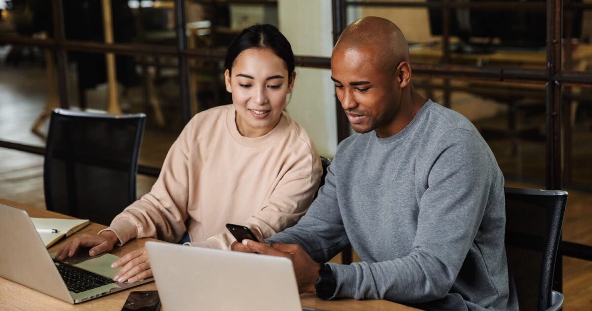 Duas pessoas sentadas à mesa, concentradas em seus laptops, trocando ideias e trabalhando juntas.