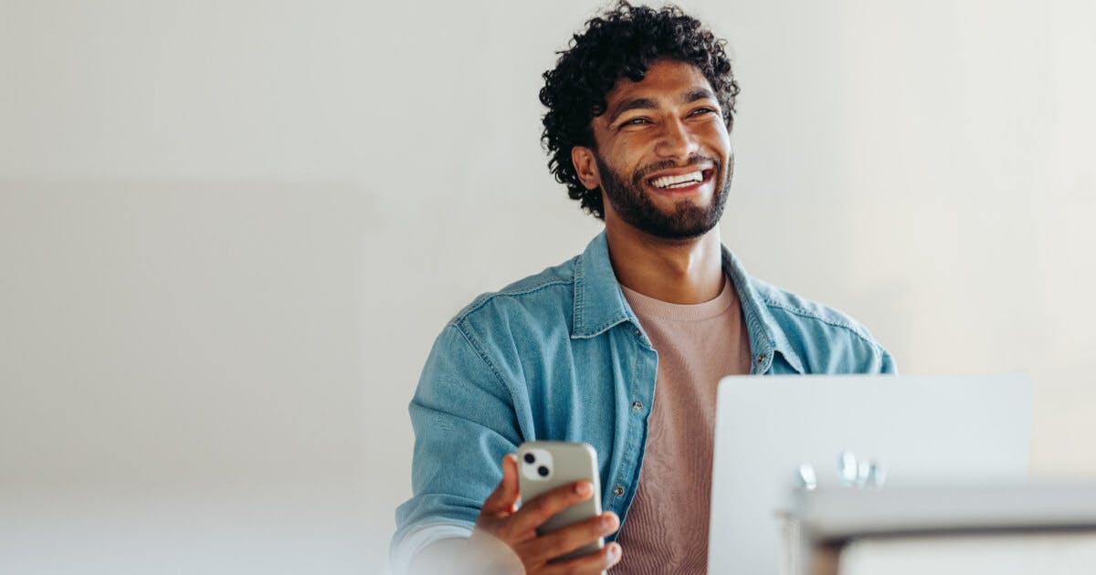 Homem sorridente utilizando um laptop e segurando um celular, transmitindo uma sensação de felicidade e produtividade.