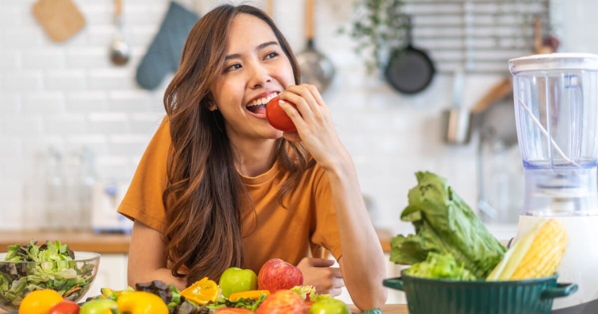 Mulher sorrindo com uma maçã na mão e frutas ao redor da mesa onde ela está escorada.